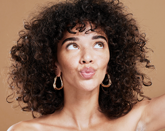Young woman with voluminous curly dark hair and gold hoop earrings against a tan backdrop, for biotin hair and beauty support