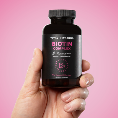 Hand holding a Vital Vitamins Biotin Complex bottle with vitamin C and hyaluronic acid against a bright pink background, for hair and nails